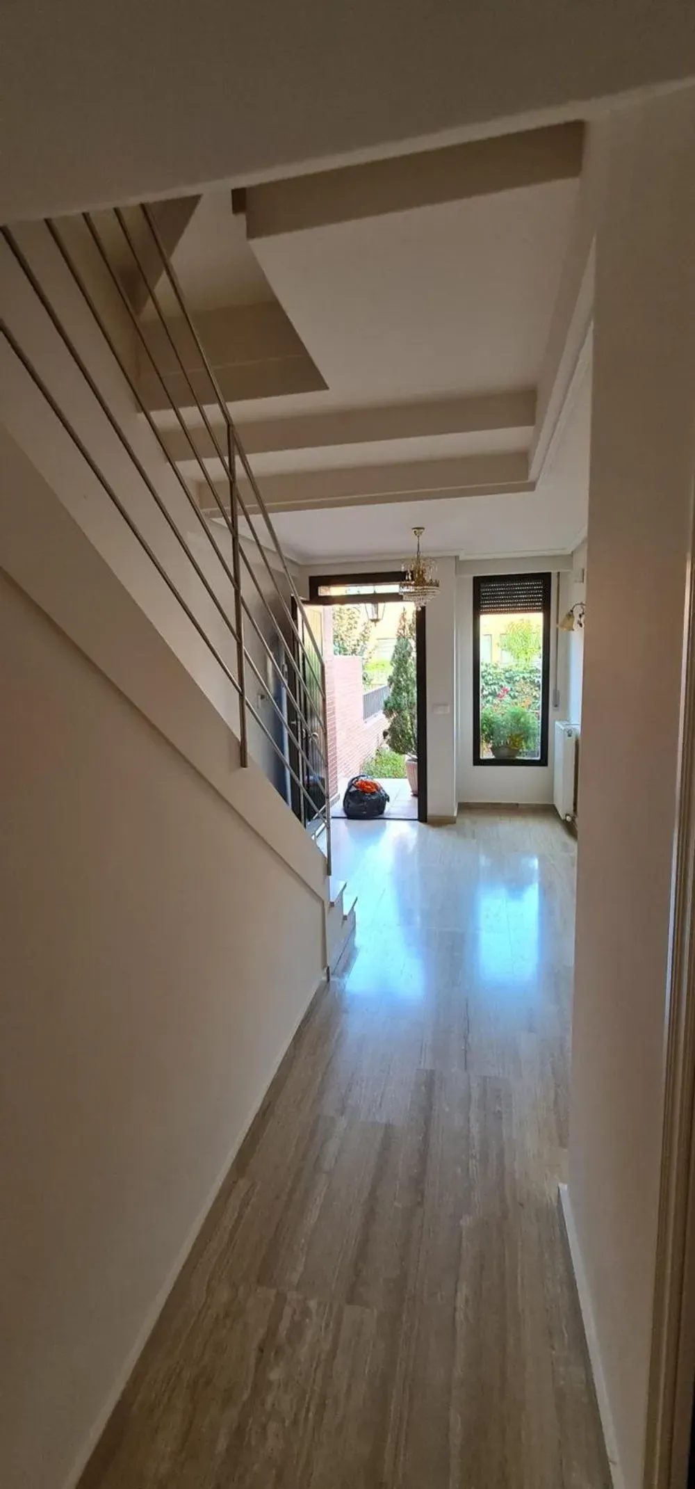 Pasillo interior con escalera, paredes blancas y suelo de madera que conduce a una puerta abierta con vista a la vegetación.