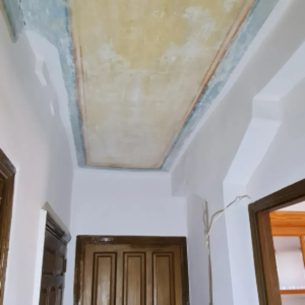 Techo de pasillo con diseño pintado en amarillo y azul. Paredes blancas y puertas de madera.