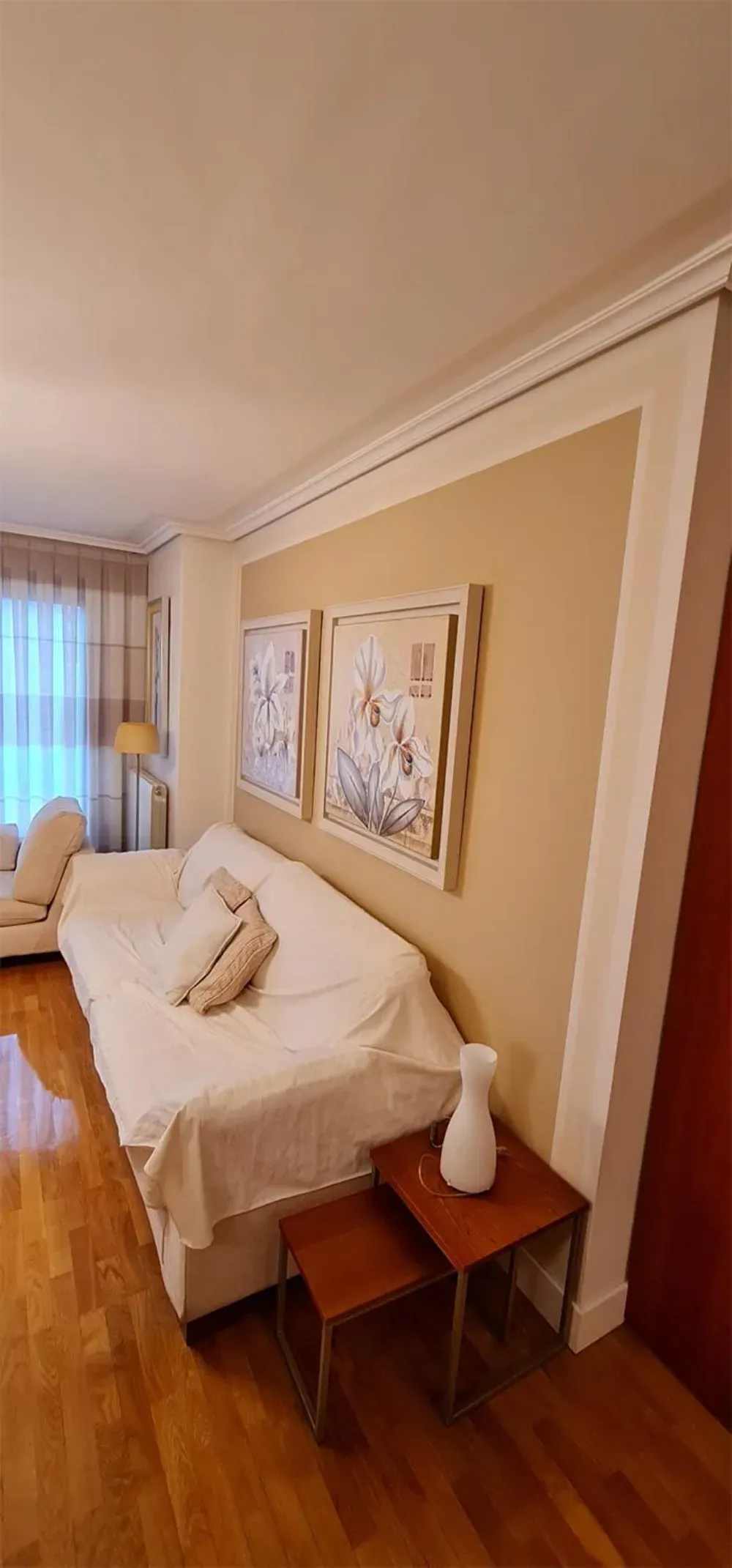 Una habitación con un sofá, dos cuadros enmarcados, dos mesas auxiliares y un jarrón sobre suelo de madera.