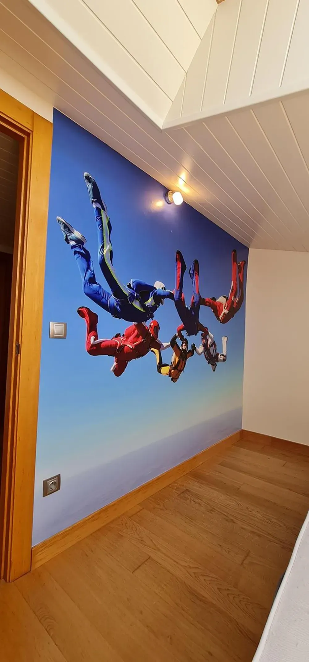 Mural de pared de paracaidistas con trajes coloridos contra un cielo azul, montado en una pared inclinada.