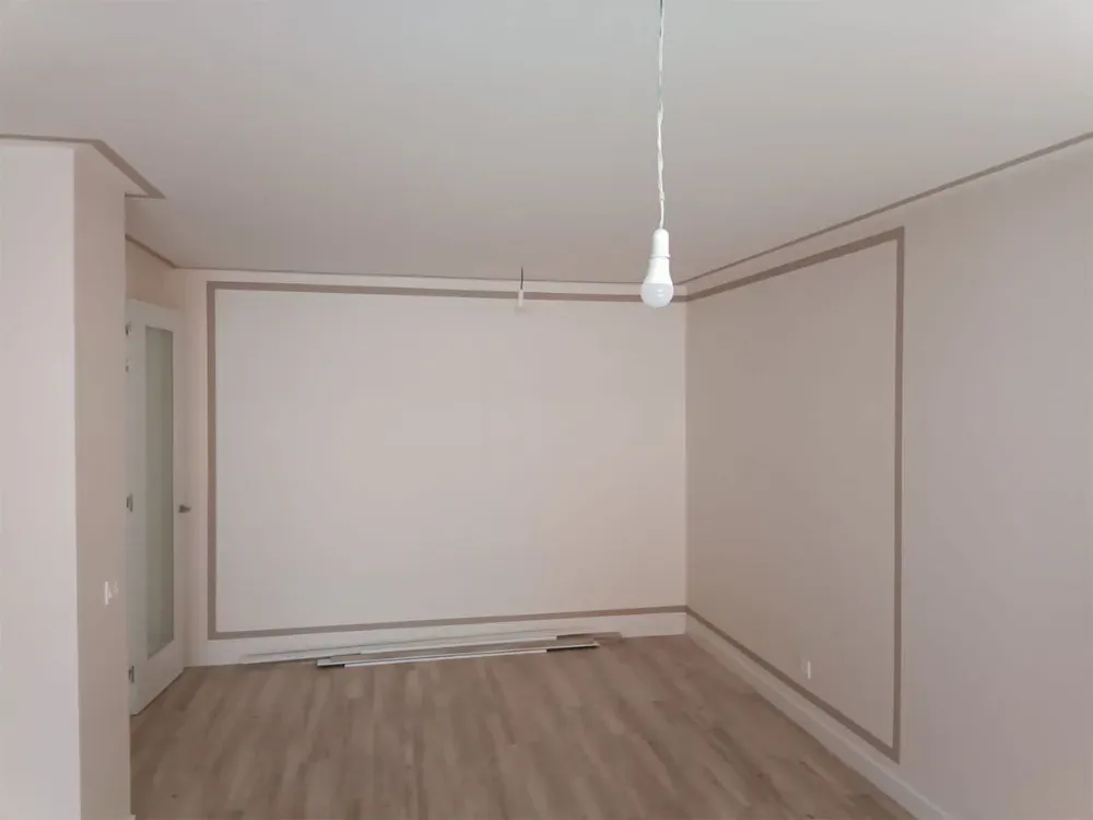 Habitación vacía con paredes blanquecinas, suelo de madera y molduras beige. Una bombilla cuelga del techo.