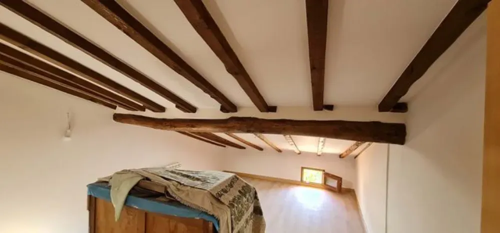 Vigas de madera se extienden sobre un techo blanco en una habitación con un armario de madera y una pequeña ventana.