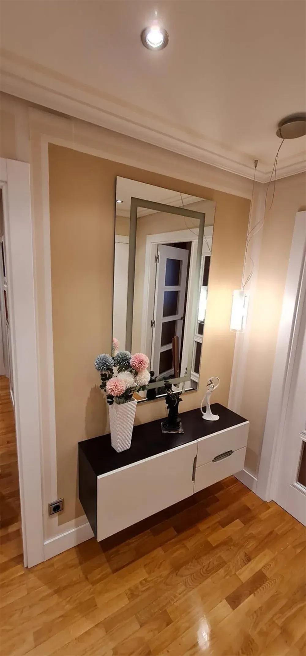 Un espejo, flores y un mueble flotante en un pasillo con suelo de madera.