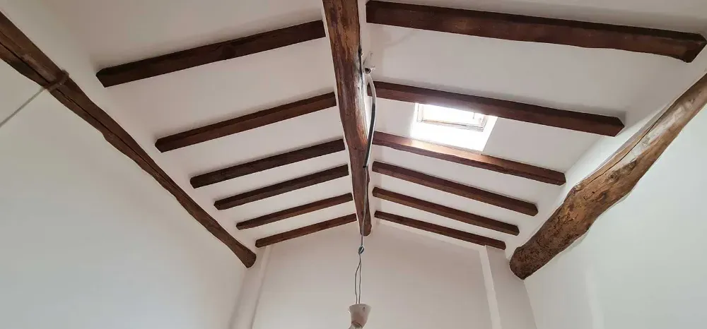 Habitación blanca con vigas de madera en el techo, un tragaluz y una lámpara colgante.