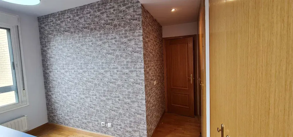 Vista interior de una habitación con paredes estampadas, una puerta de madera y una ventana parcialmente visible.