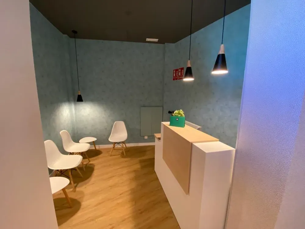 Sala de espera con sillas blancas, paredes azul pálido, piso de madera y mostrador de recepción.