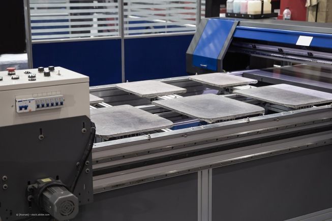 Großformatdrucker mit mehreren flachen, grauen Objekten auf einem Metallgitter, wahrscheinlich in einer Fabrik.