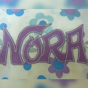 El nombre Nora está bordado en morado sobre un fondo blanco.