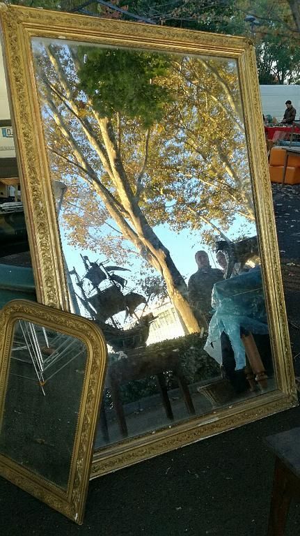 Miroir à cadre doré reflétant des arbres et des passants à l'extérieur d'un marché.
