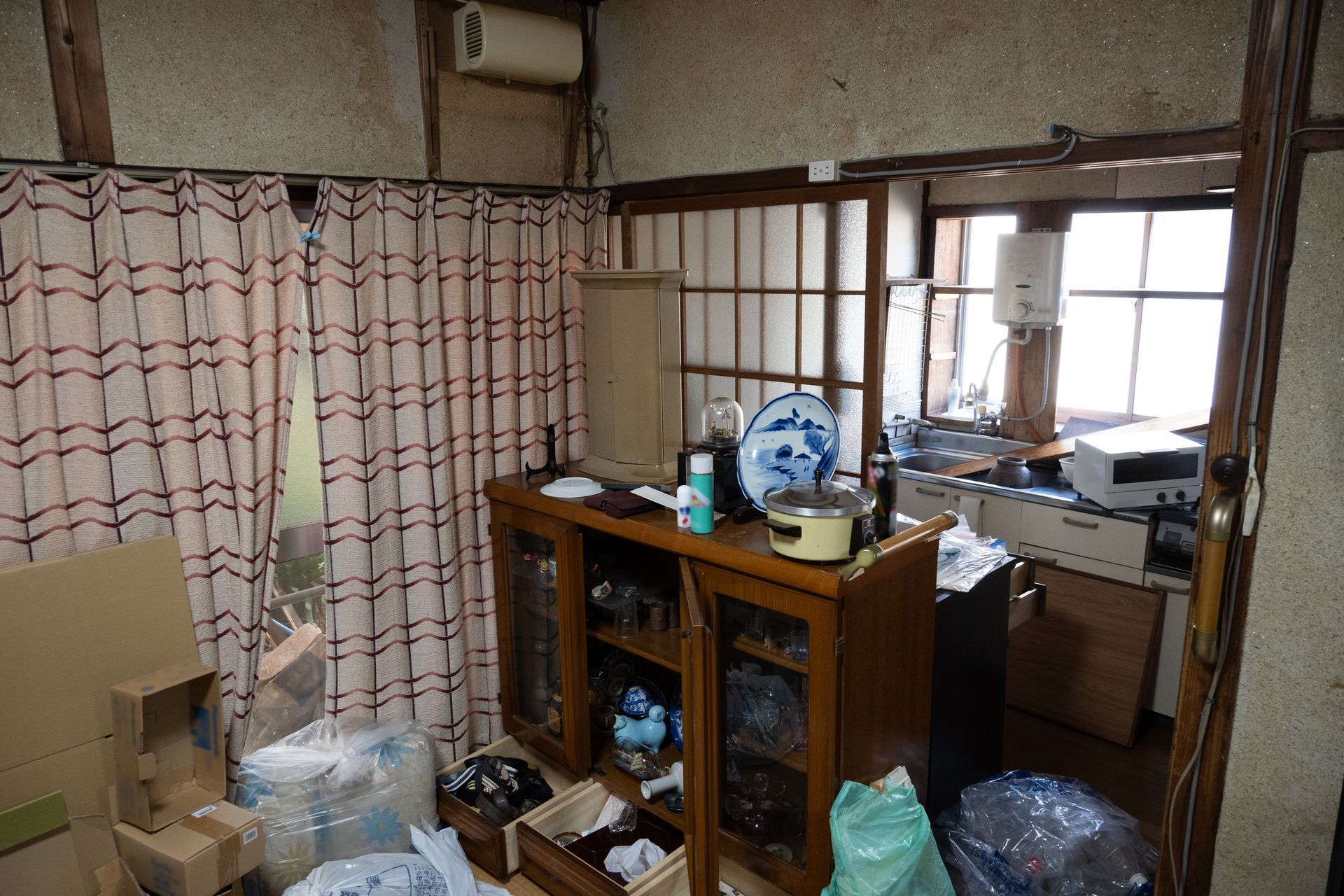 Pièce encombrée avec des rideaux aux fenêtres, un buffet en bois, des cartons empilés et des objets ménagers éparpillés çà et là.