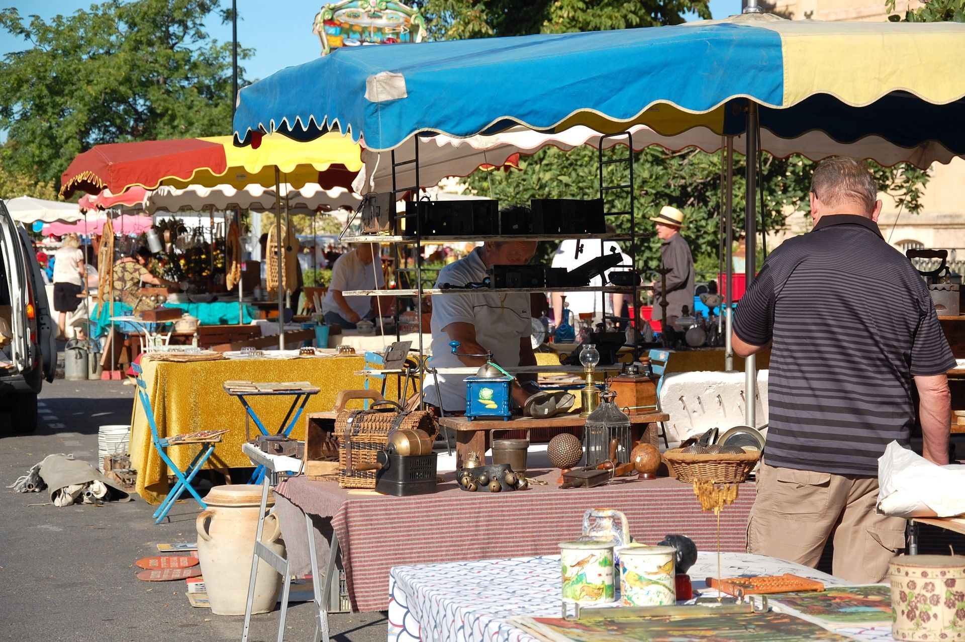 Étal de marché en plein air avec un auvent bleu, des tables remplies de marchandises et une personne qui regarde à proximité