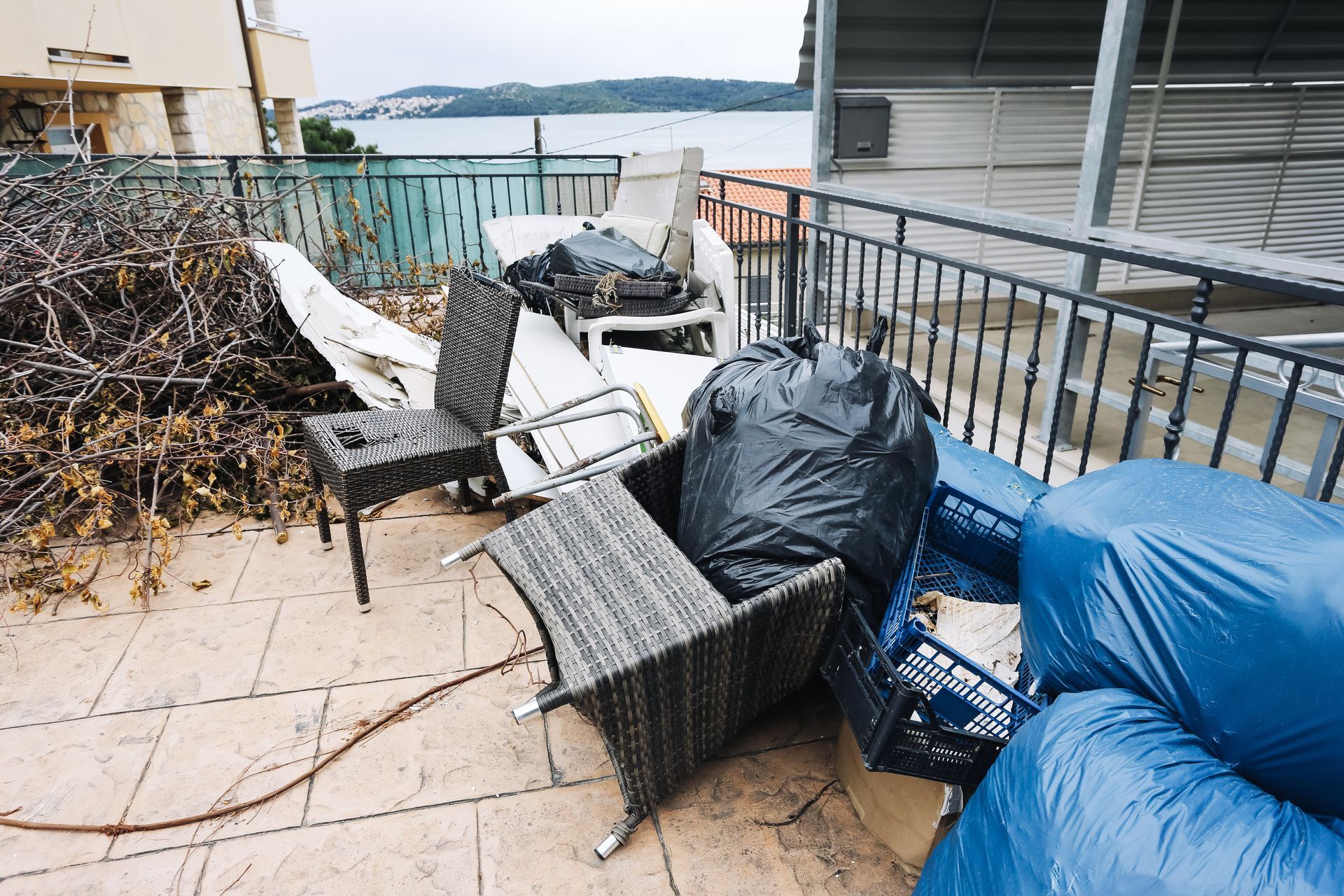 Terrasse avec chaises en osier renversées, coussins bleus et débris près d'une rambarde au bord de l'eau.