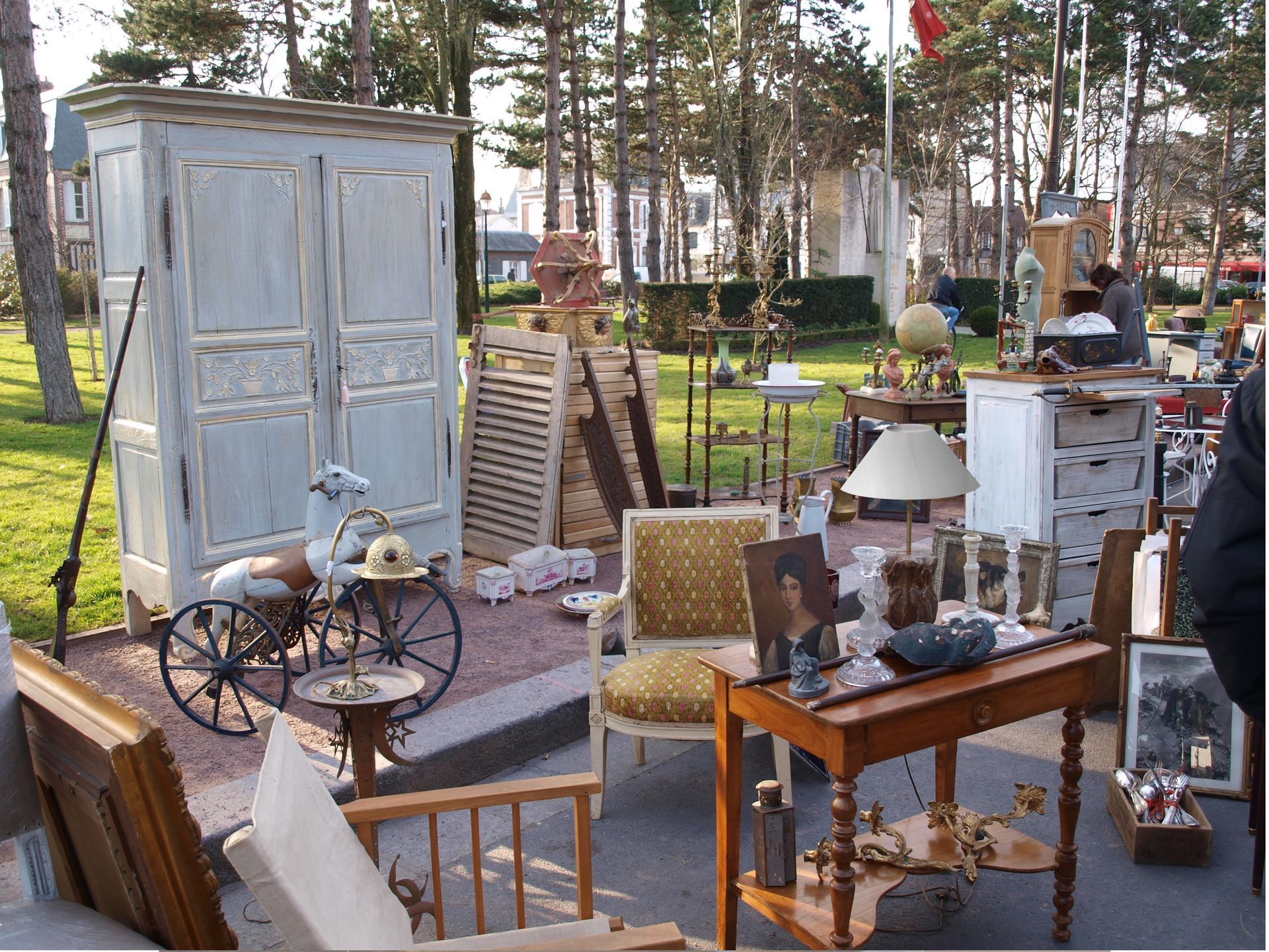 Marché aux puces en plein air à proximité d'arbres avec des meubles vintage, une armoire blanche, un fauteuil et des petites tables.