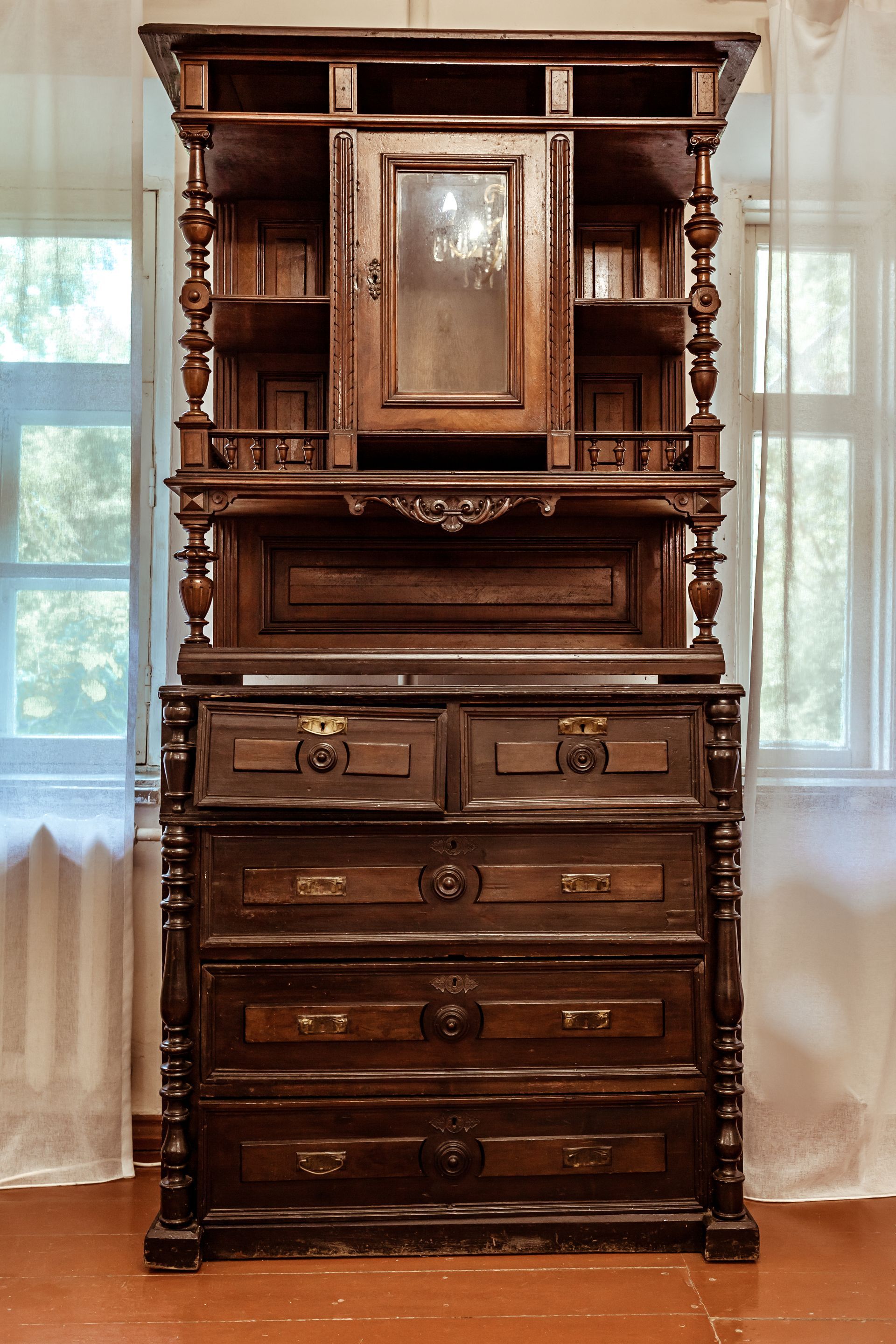 Armoire ancienne en bois sculptée avec placard vitré au centre et étagères sur les côtés, placée devant des fenêtres lumineuses.