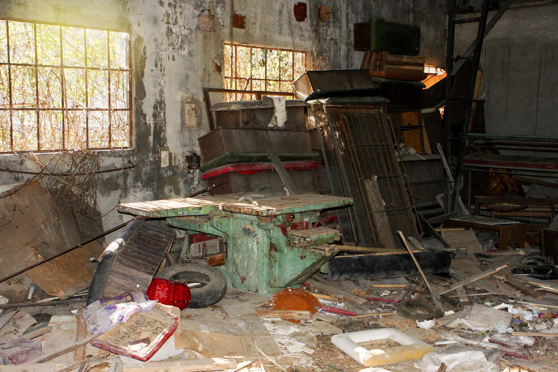 Pièce abandonnée et encombrée de débris, avec des meubles cassés, des fenêtres sales, des déchets et des morceaux de verre éparpillés.