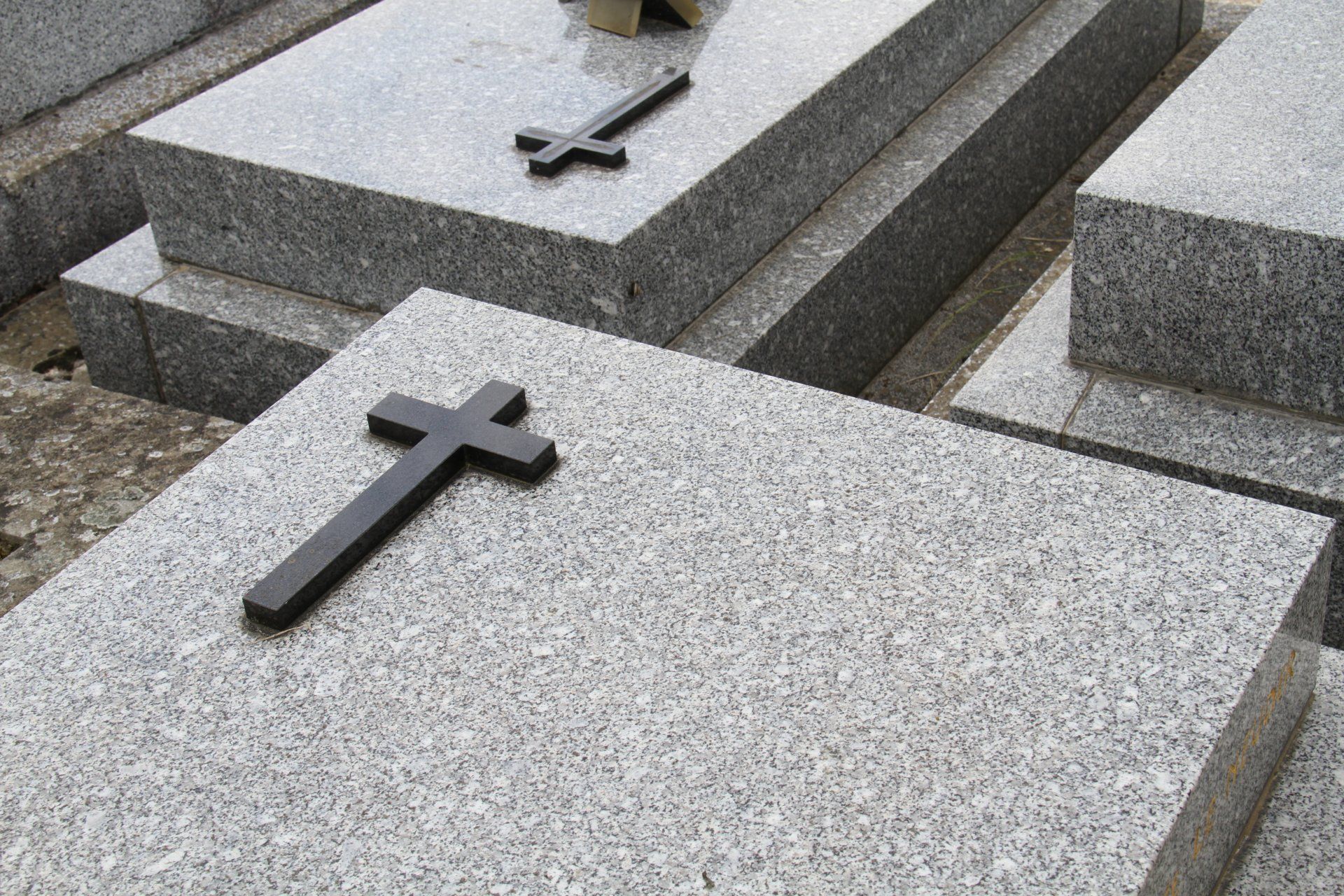 Des pierres tombales en granit gris surmontées de croix sombres, dans un cimetière.