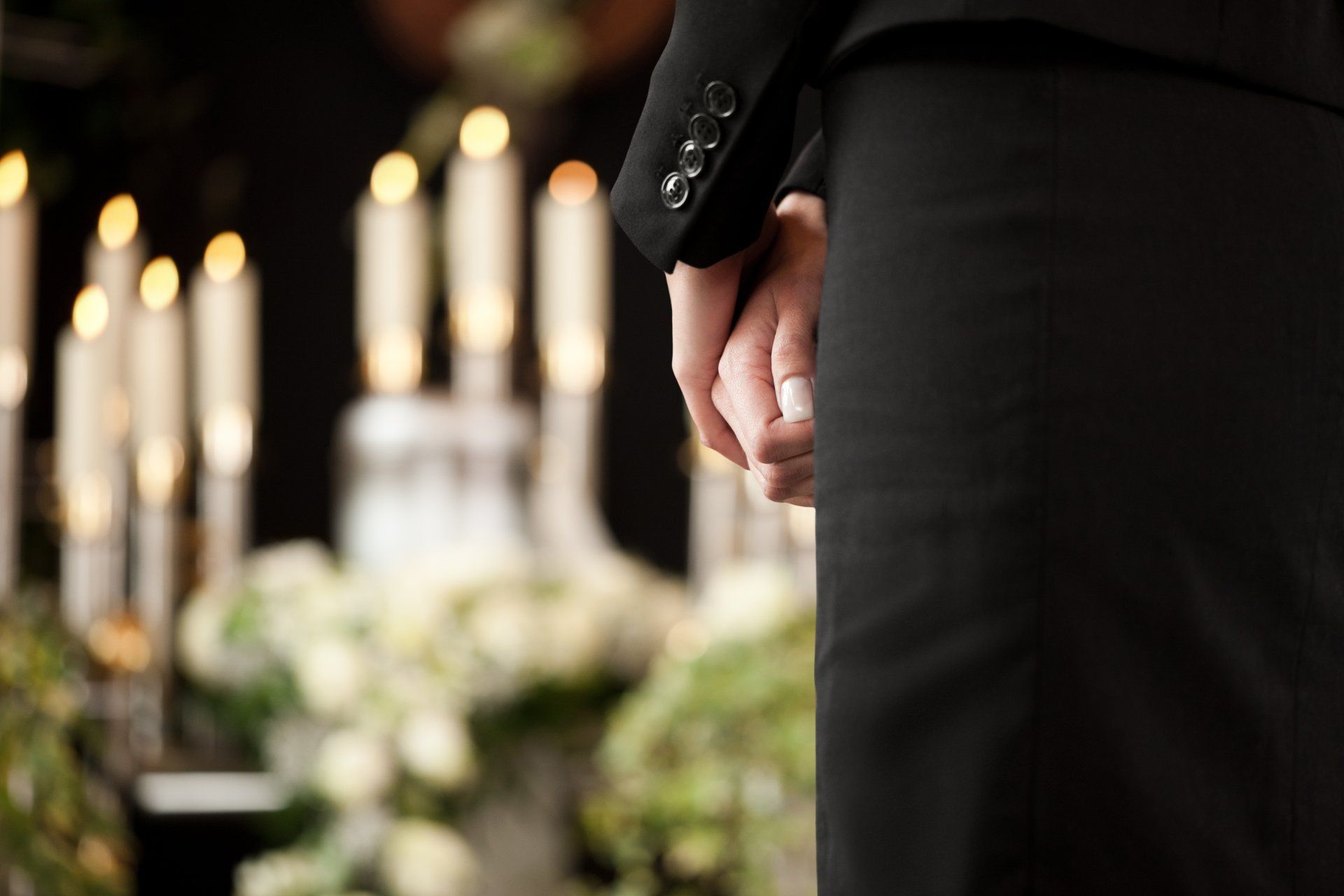 Personne vêtue de noir, debout lors d'un enterrement ; mains jointes, bougies et fleurs en arrière-plan.