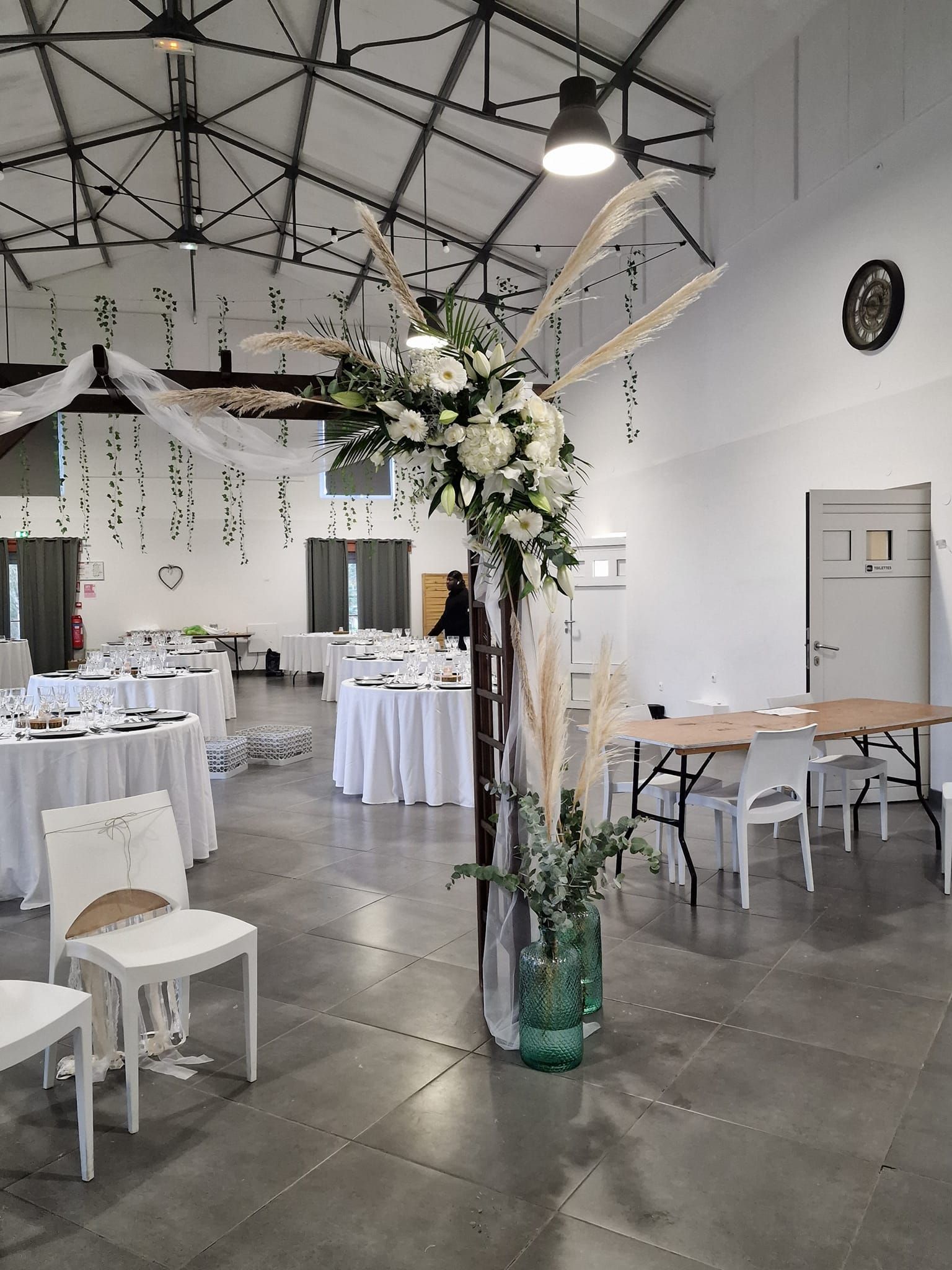 Salle de réception avec décoration florale et tables dressées pour les invités. Les tons blancs, verts et bruns dominent l'espace.