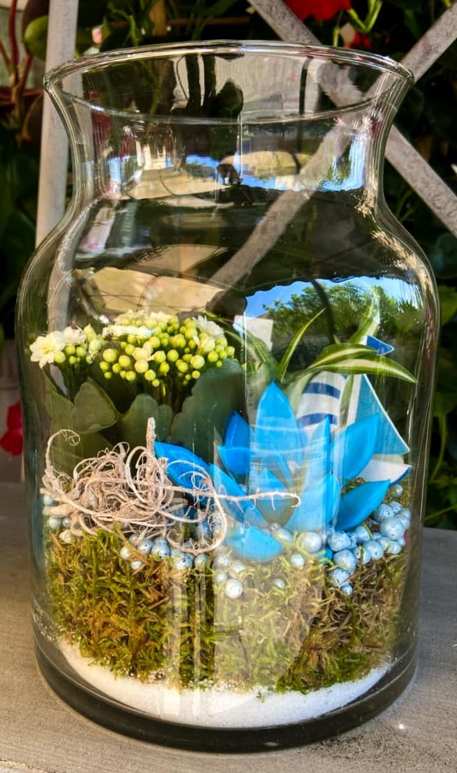 Terrarium en verre avec mousse, accents bleus et détails de fausses plantes.