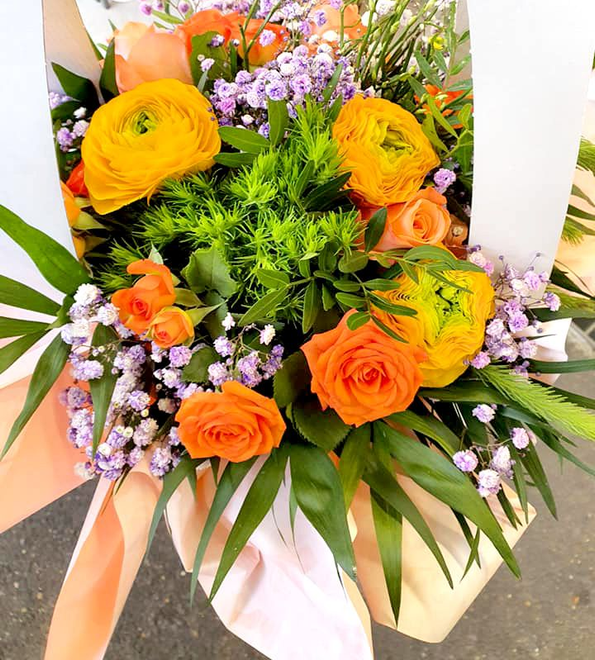 Bouquet de fleurs orange et jaunes vibrantes, dont des roses et des renoncules, entouré de verdure et de gypsophile.