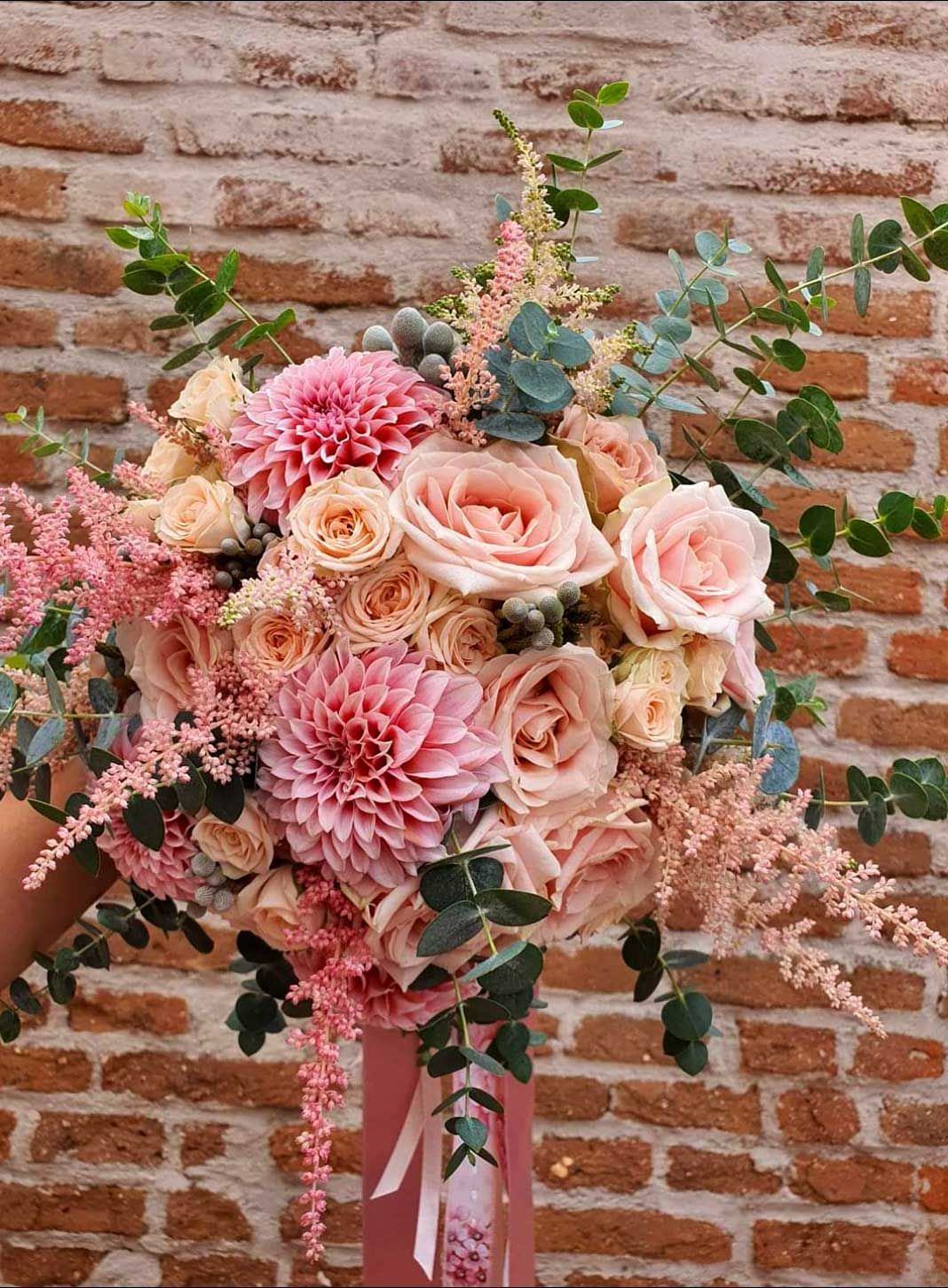 Bouquet de fleurs roses contre un mur de briques, comprenant des dahlias, des roses et de la verdure.