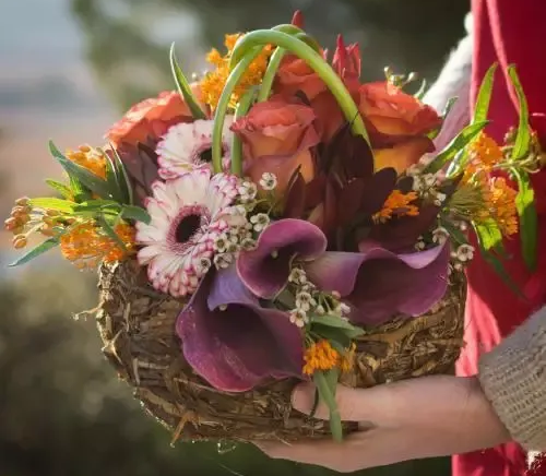 Personne tenant un panier de fleurs colorées : roses, lys calla et marguerites gerberas.
