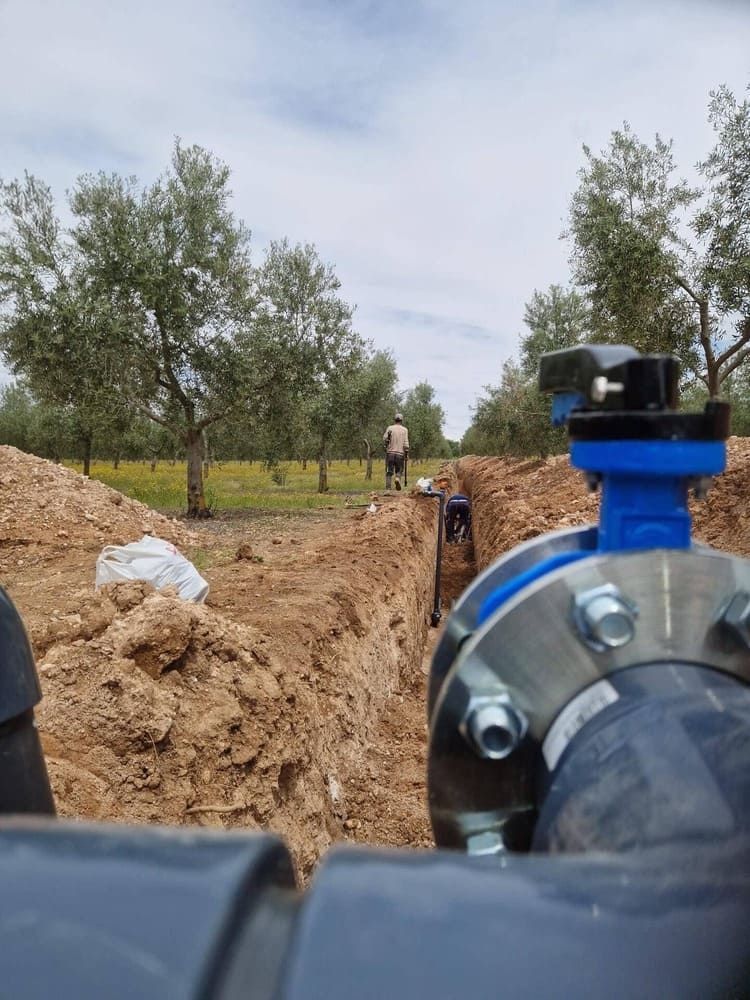 Zanja con una válvula azul de tubería en primer plano, montones de tierra y árboles al fondo.