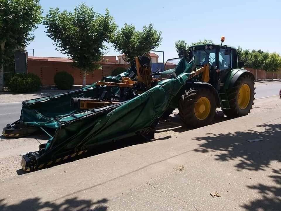 Máquina agrícola verde con un apero plegado, estacionada en una calle soleada cerca de edificios y árboles