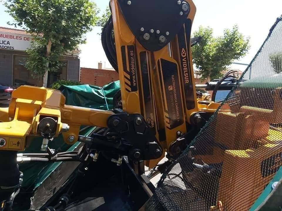 Brazo de excavadora amarillo trabajando junto a una valla de malla al aire libre, con árboles y maquinaria de construcción al fondo