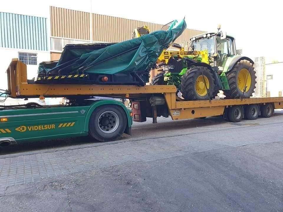 Máquina agrícola verde sobre un remolque de plataforma junto a un tractor en una calle