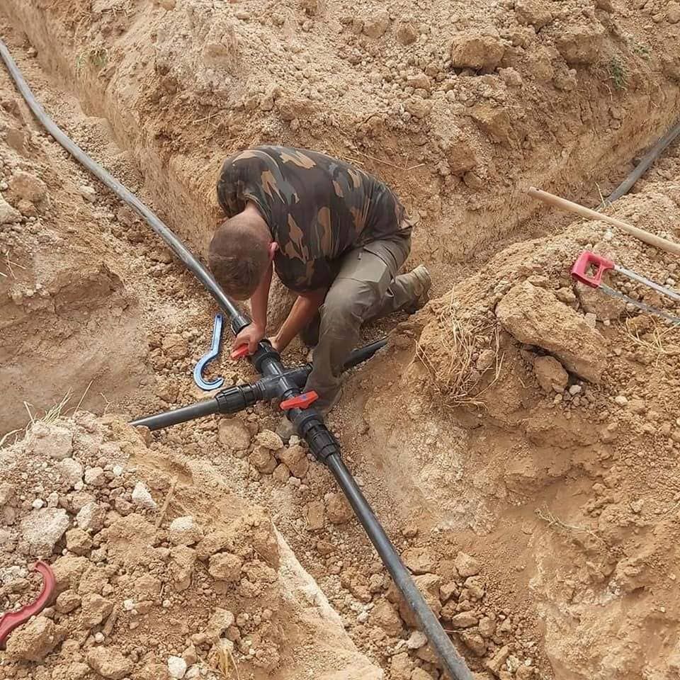 Un trabajador conecta tuberías negras en una zanja de tierra con herramientas rojas