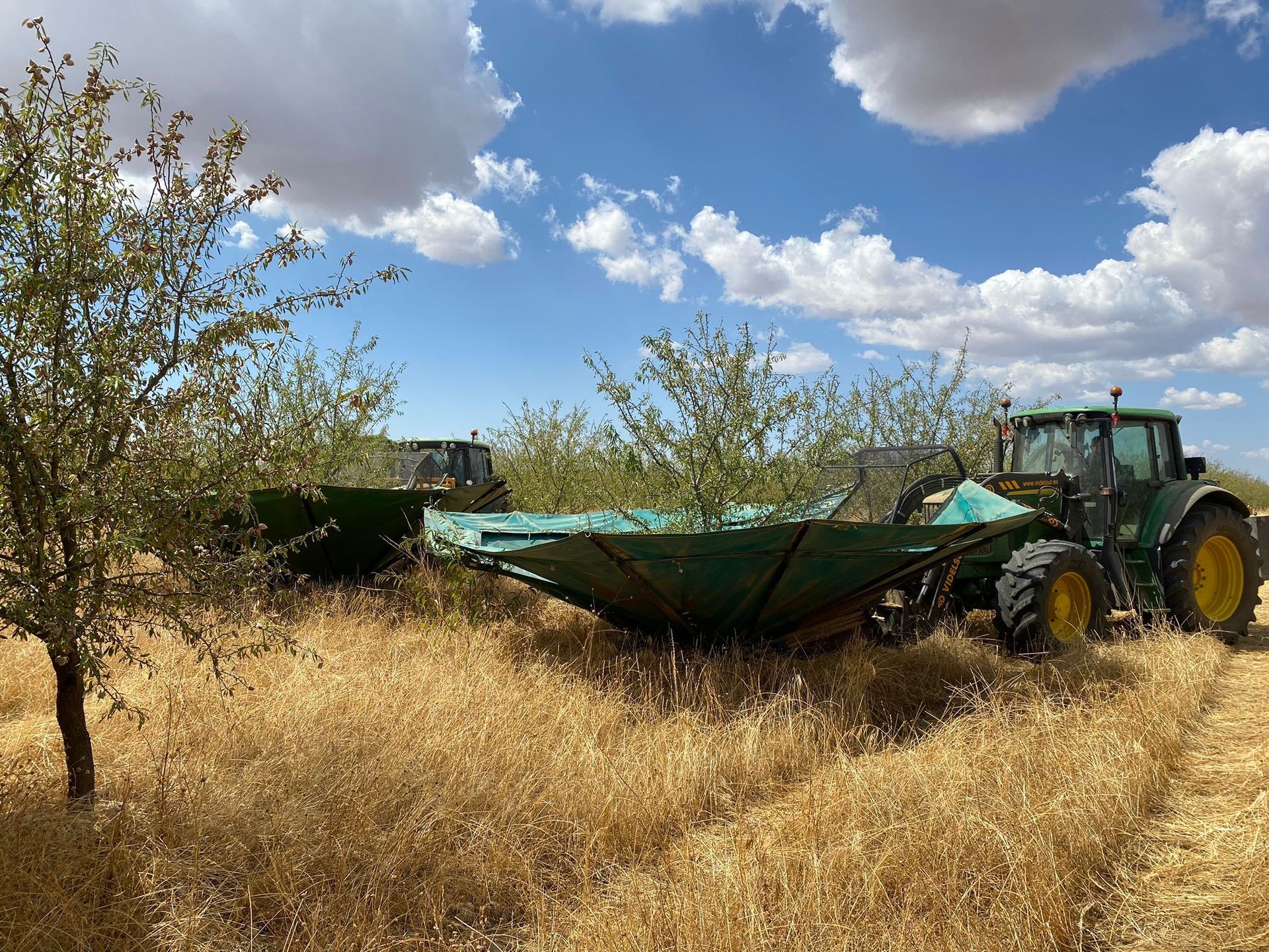 Un tractor verde arrastra un rastrillo de heno por un campo seco bajo un cielo parcialmente nublado