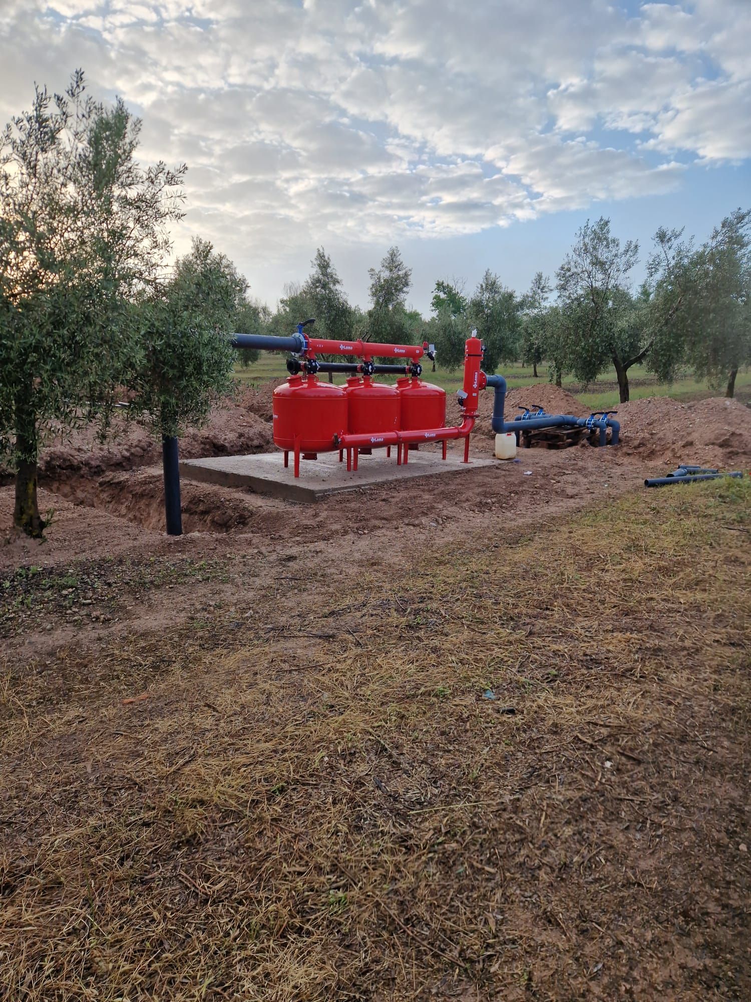 Máquina industrial roja en un claro de tierra rodeado de árboles, bajo un cielo nublado