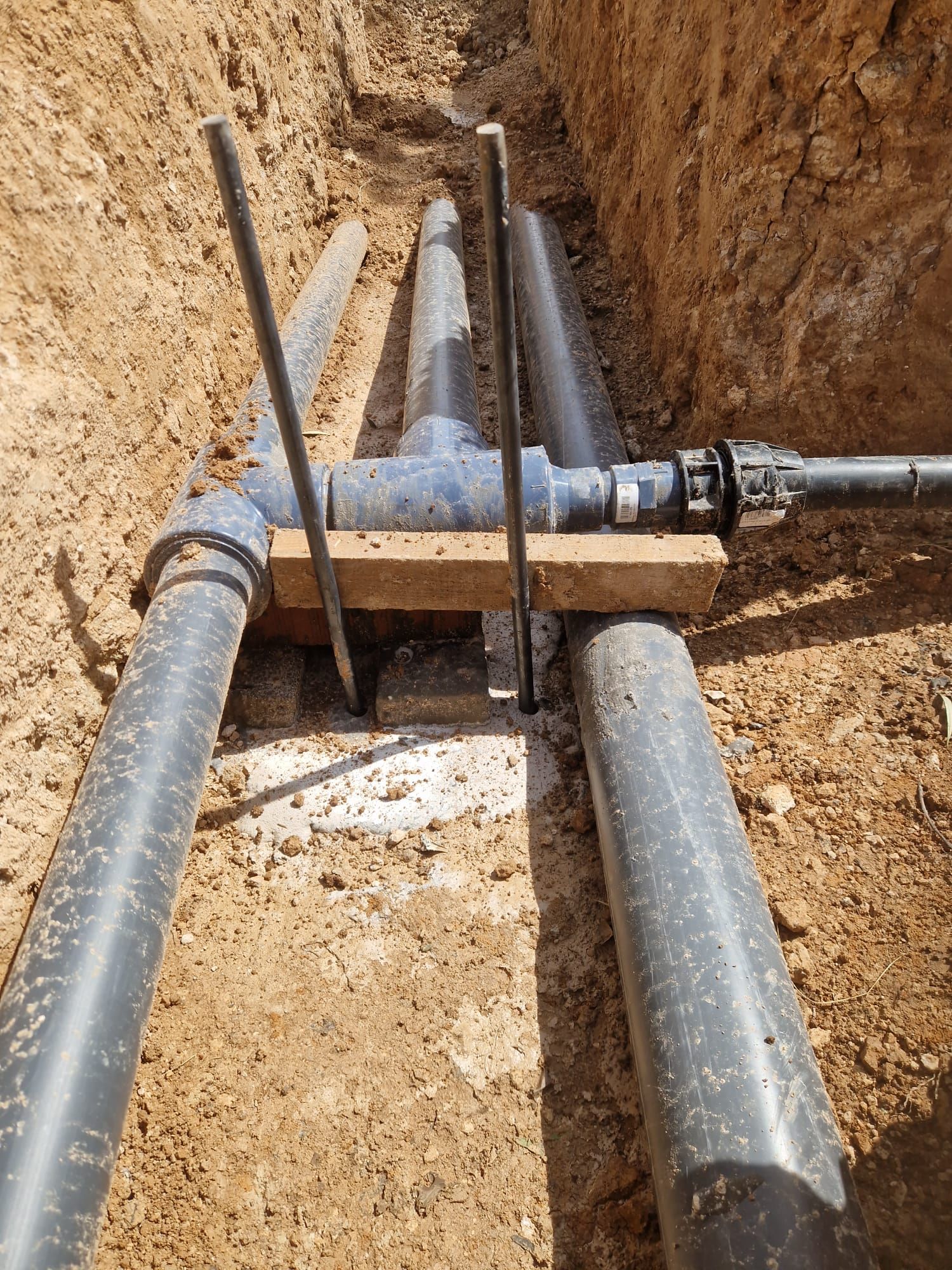 Dos tuberías negras de servicio conectadas por un tubo transversal blanco en una zanja de tierra