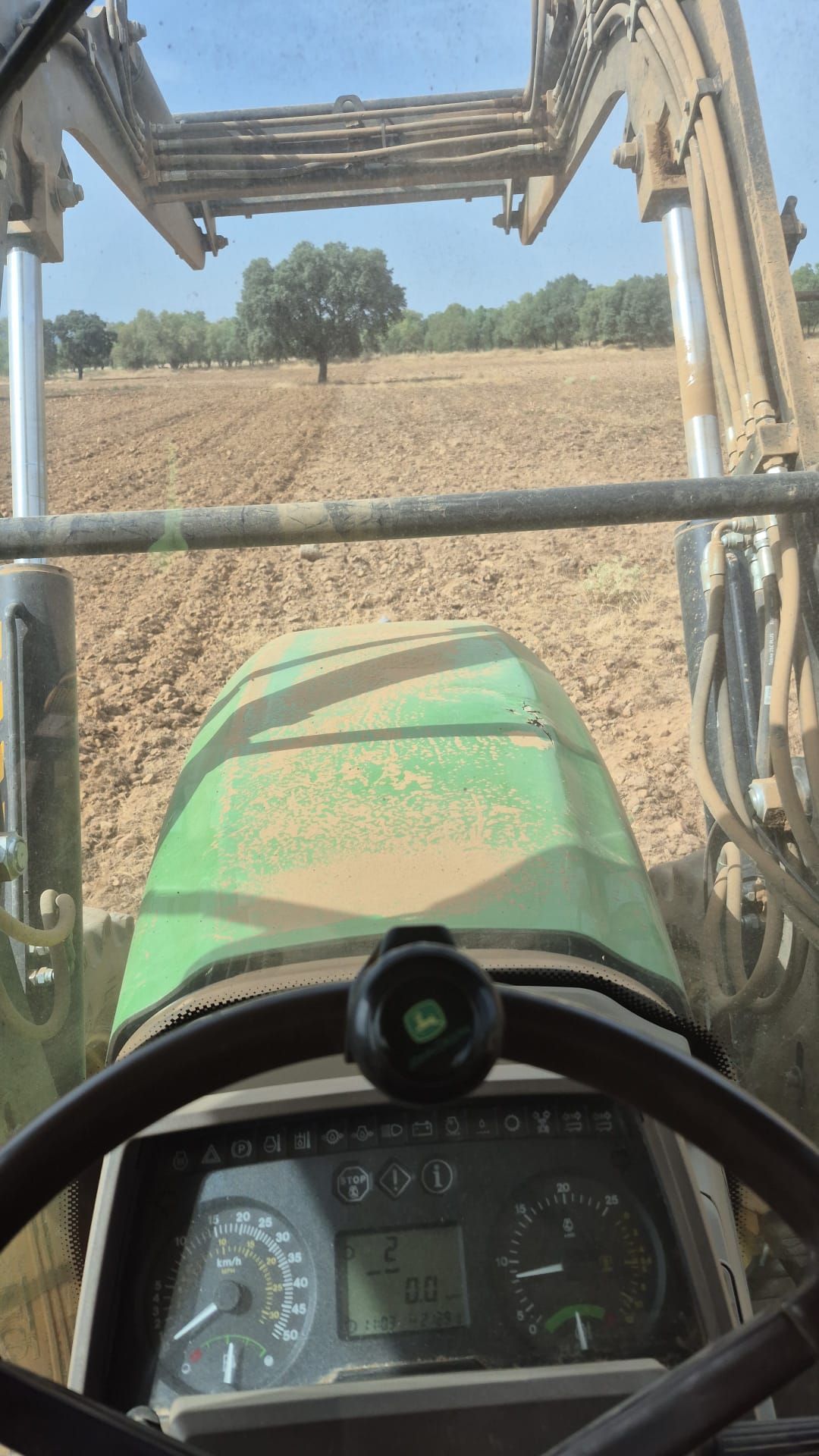 Vista desde la cabina de un tractor sobre un campo arado, con el capó verde y la pala cargadora delantera a la vista.