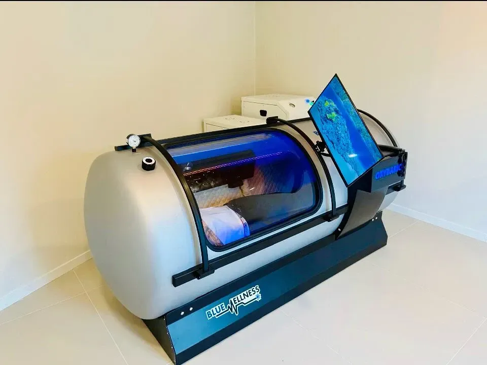 Una persona dentro de una cámara hiperbárica plateada con un interior azul, una pantalla que muestra un video