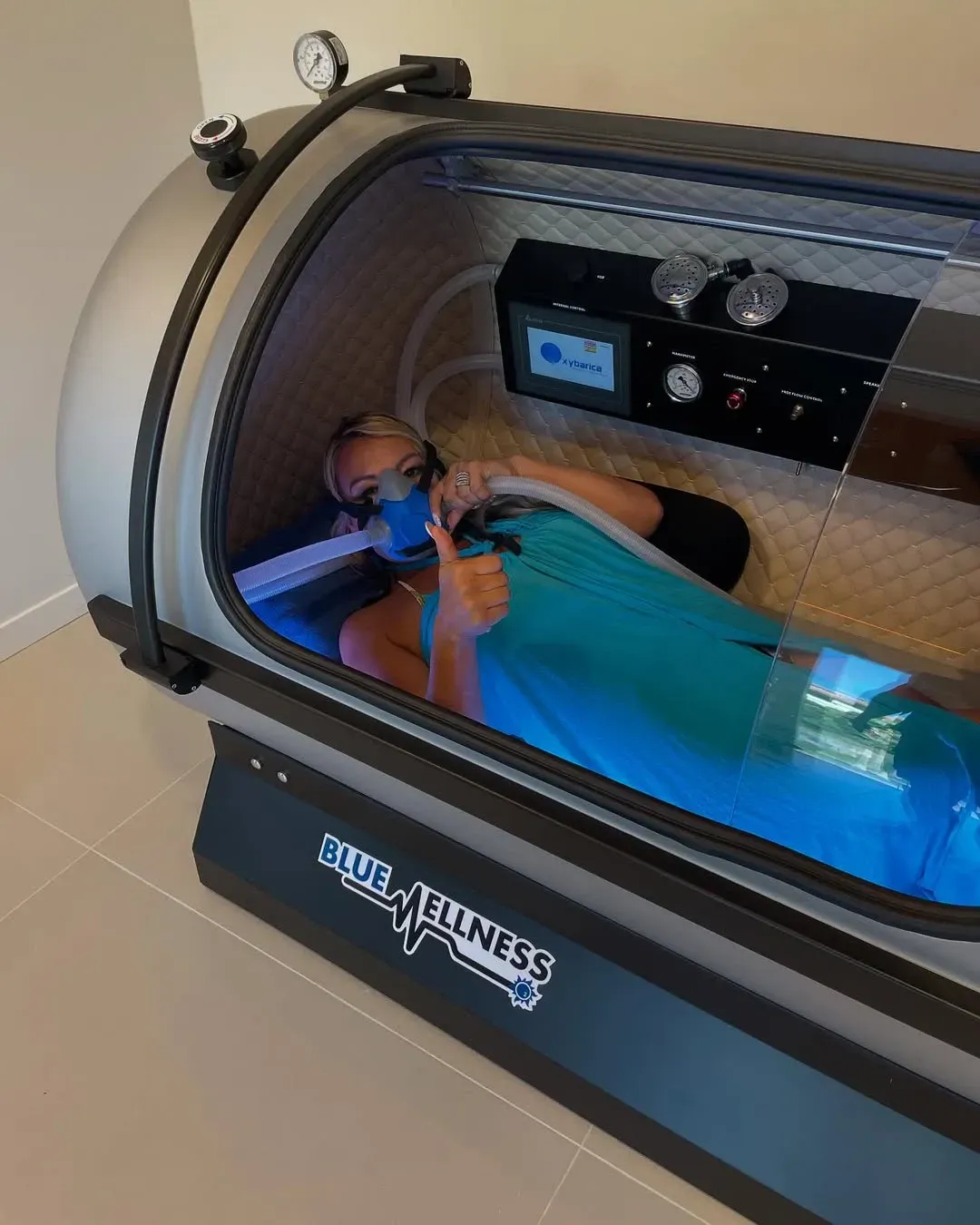 Mujer dentro de una cámara hiperbárica con máscara de oxígeno, haciendo un gesto de aprobación con el pulgar.