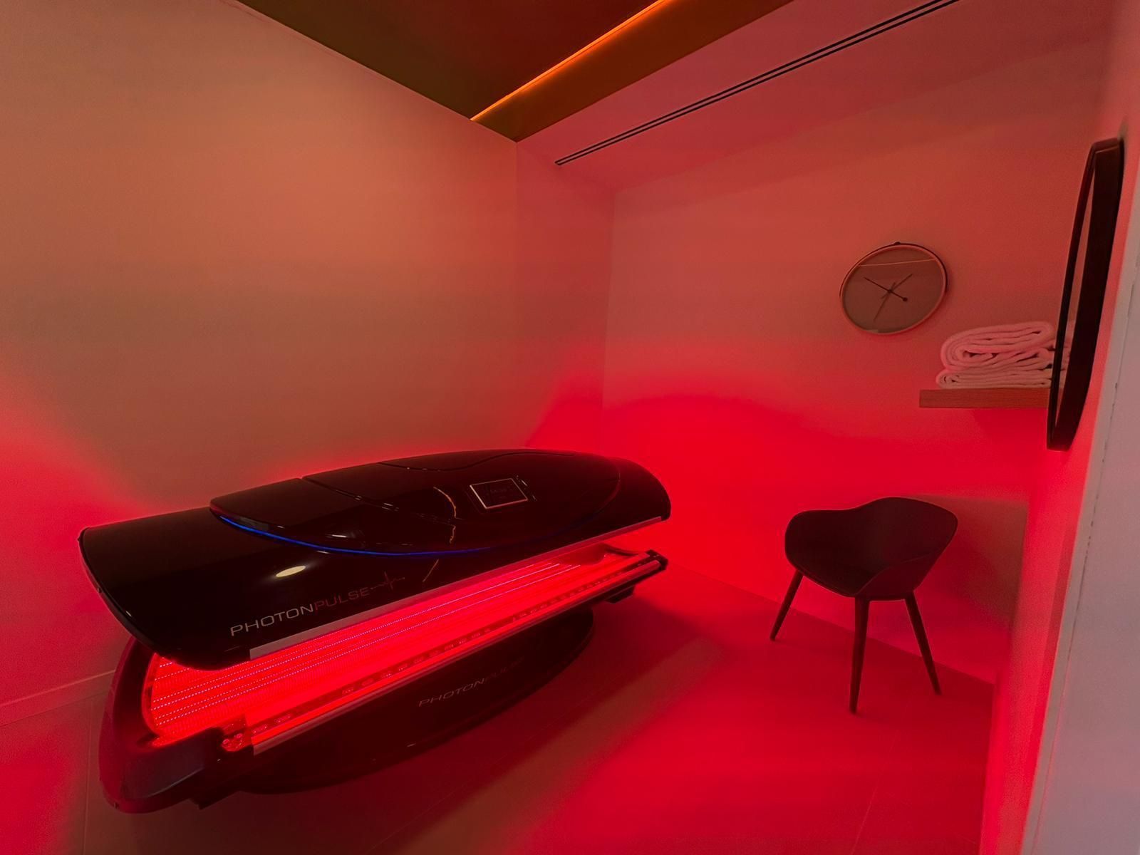 Cama de terapia de luz roja en una habitación blanca con una pequeña silla y un reloj.