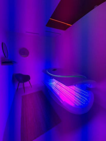 Habitación con una bañera de aspecto futurista iluminada con luces moradas y rosas. Una pequeña silla y espejos en la pared.