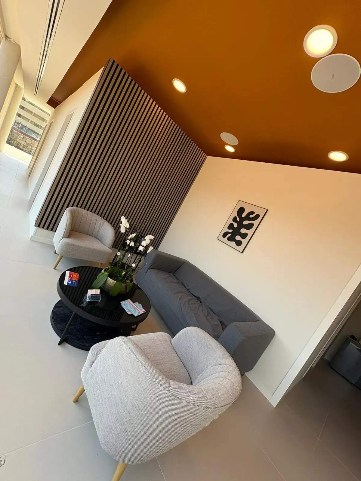 Moderna sala de espera con sofá gris, sillas mullidas, mesa de café redonda, arte y techo naranja.