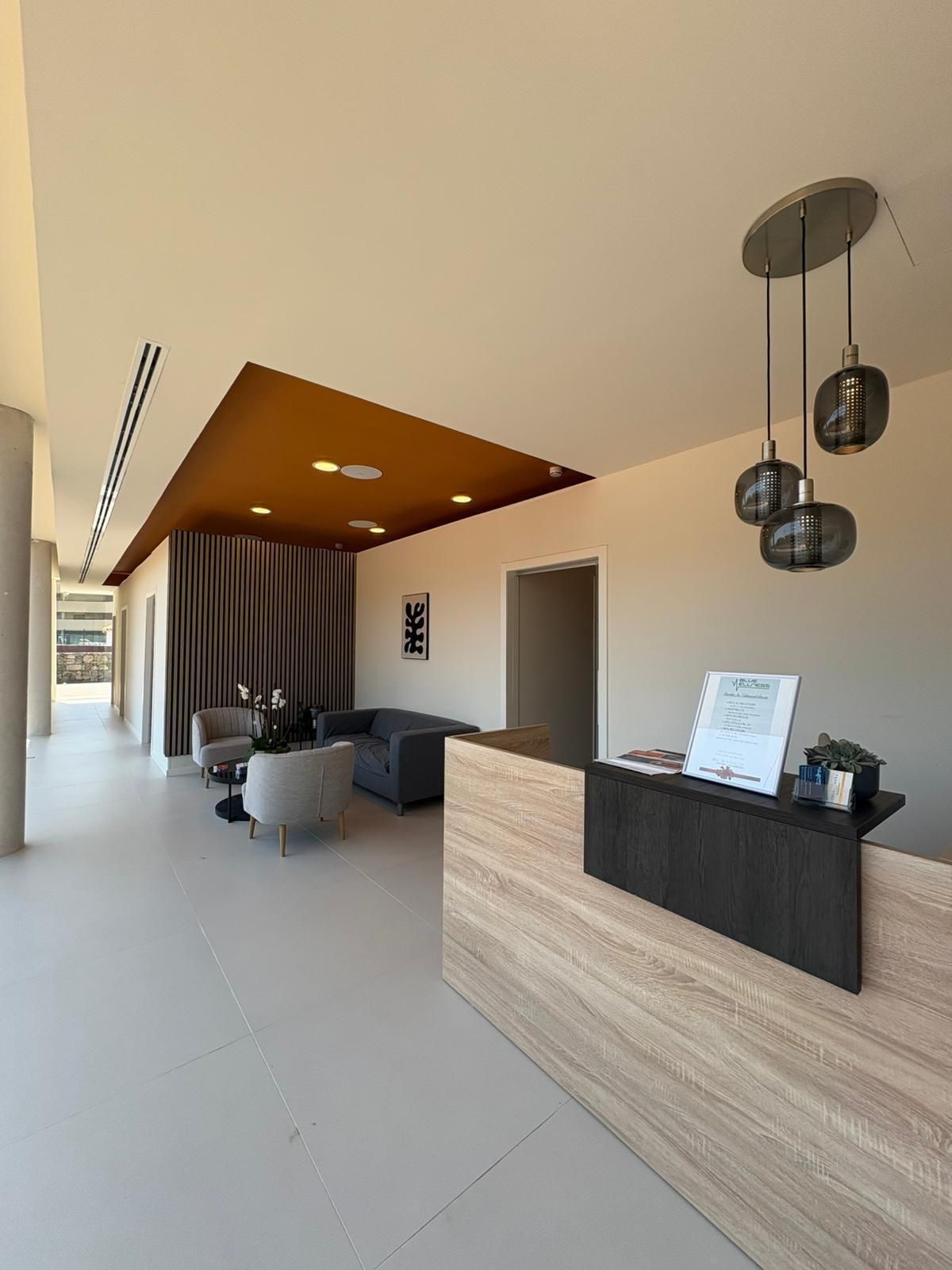 Área de recepción con escritorio de madera clara, zona de estar y lámparas colgantes.