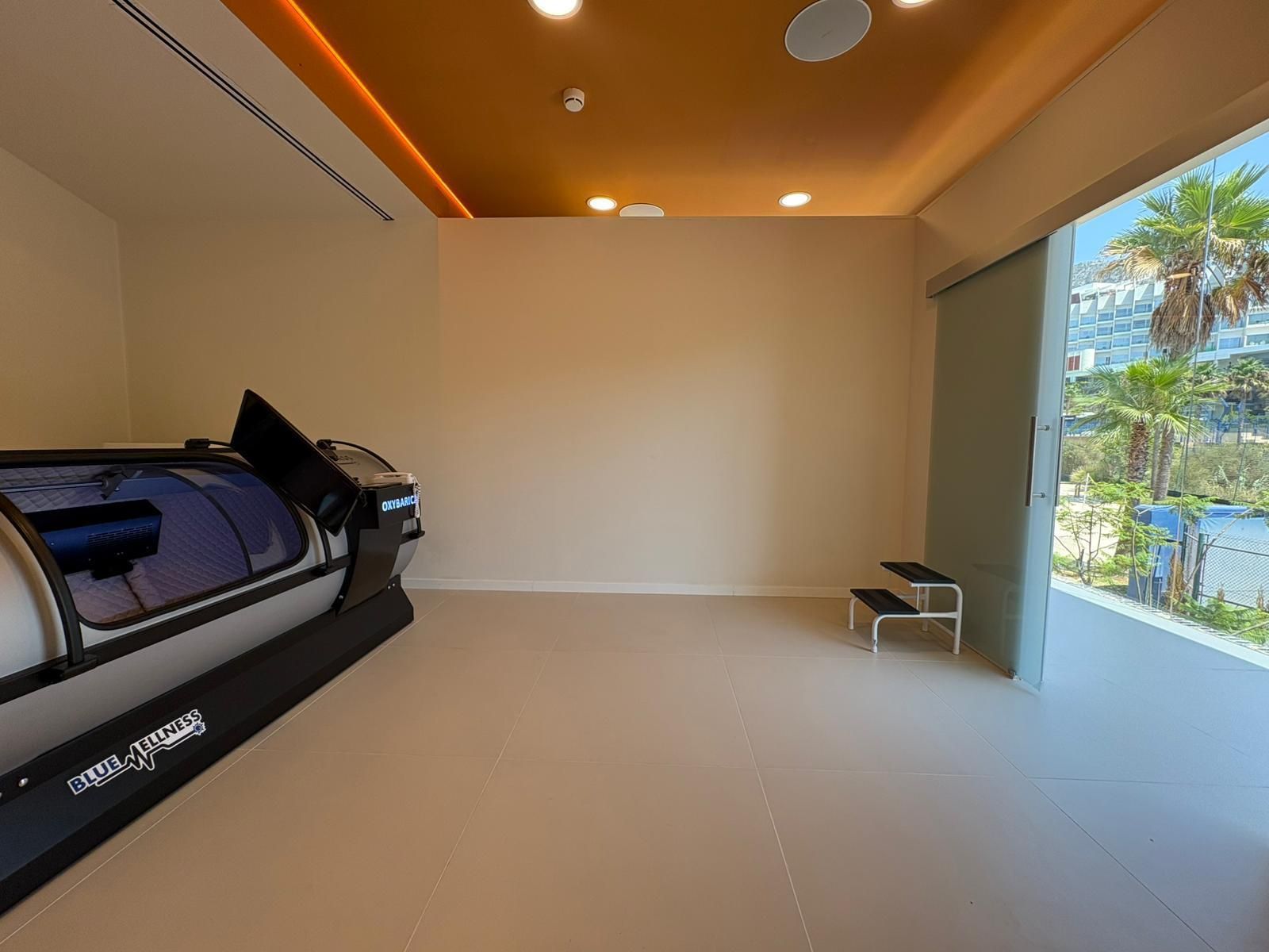 Habitación con cámara hiperbárica. Paredes y suelo beige, techo naranja y una puerta abierta a un exterior soleado.