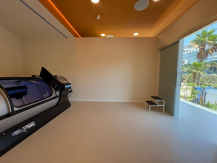 Una habitación vacía con una cámara hiperbárica a la izquierda y una puerta a un balcón soleado.