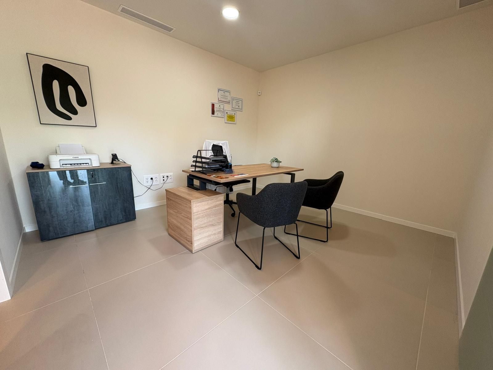 Una pequeña oficina con un escritorio, dos sillas, un armario, obras de arte y una impresora. Paredes y suelo beige.