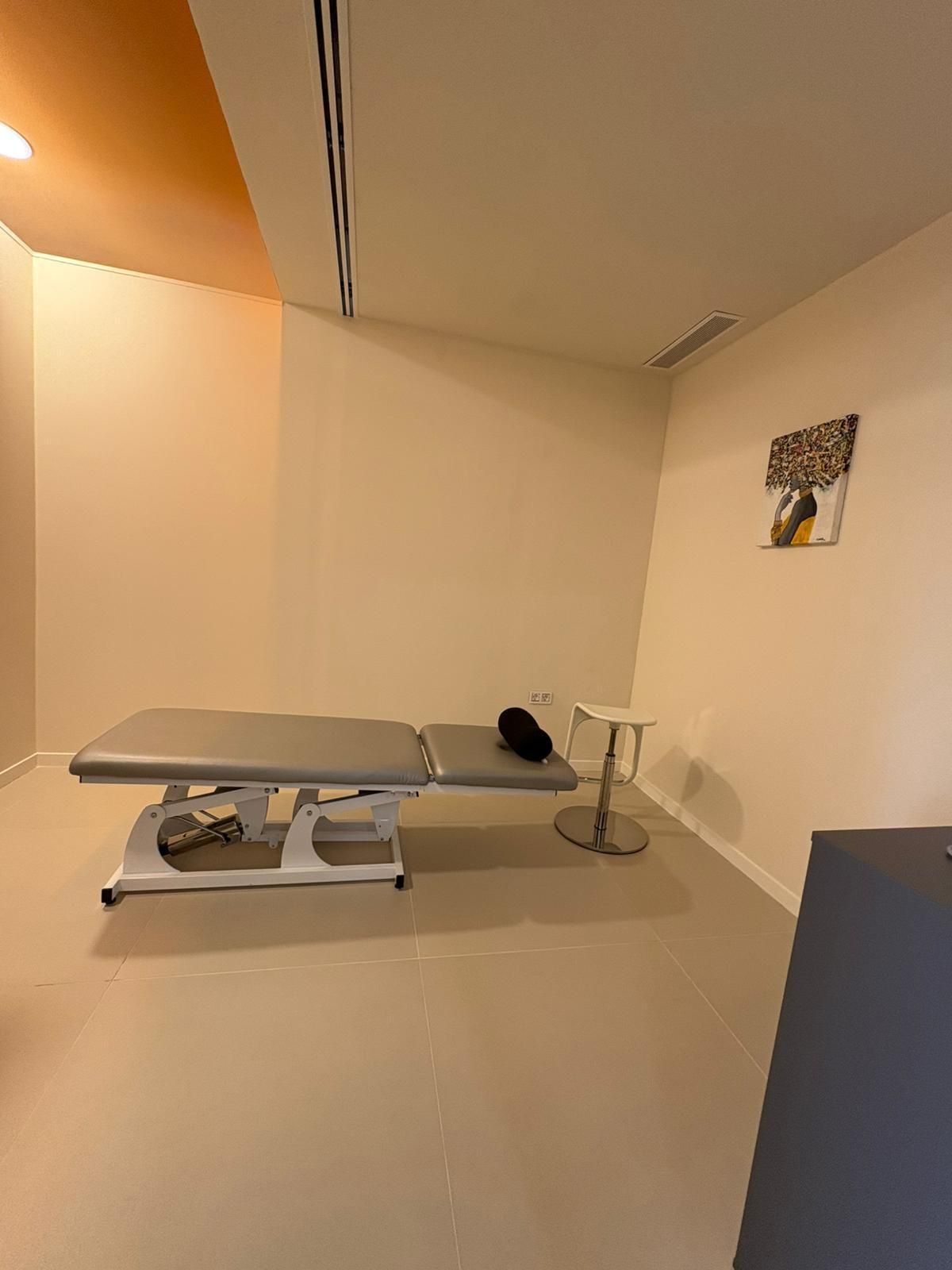 Sala de reconocimiento con camilla de masaje, taburete y obra de arte en la pared. Tonos beige y blanco.