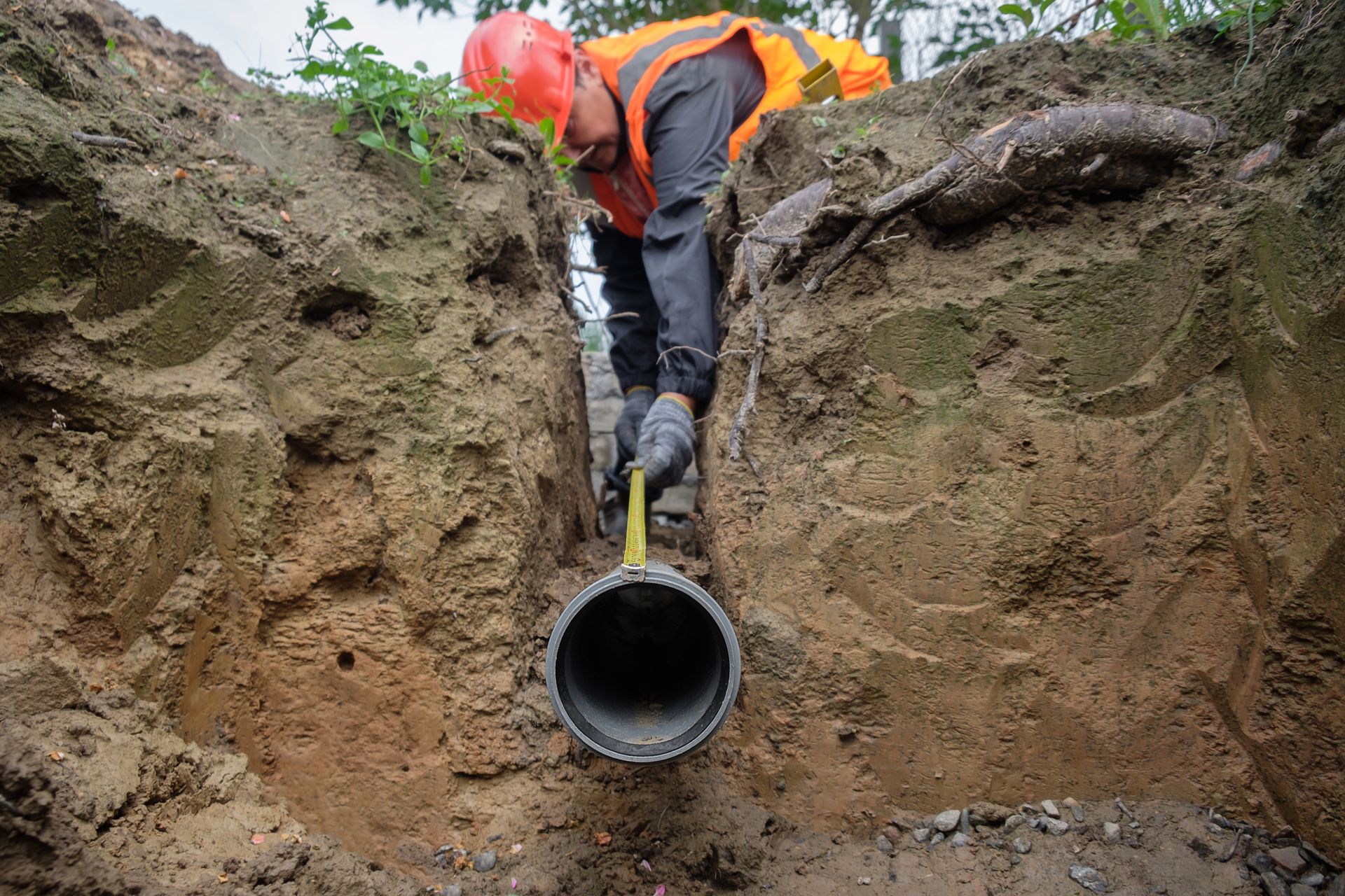 Un ouvrier en tenue de sécurité orange se tient dans une tranchée au-dessus de l'ouverture d'une grande canalisation souterraine.