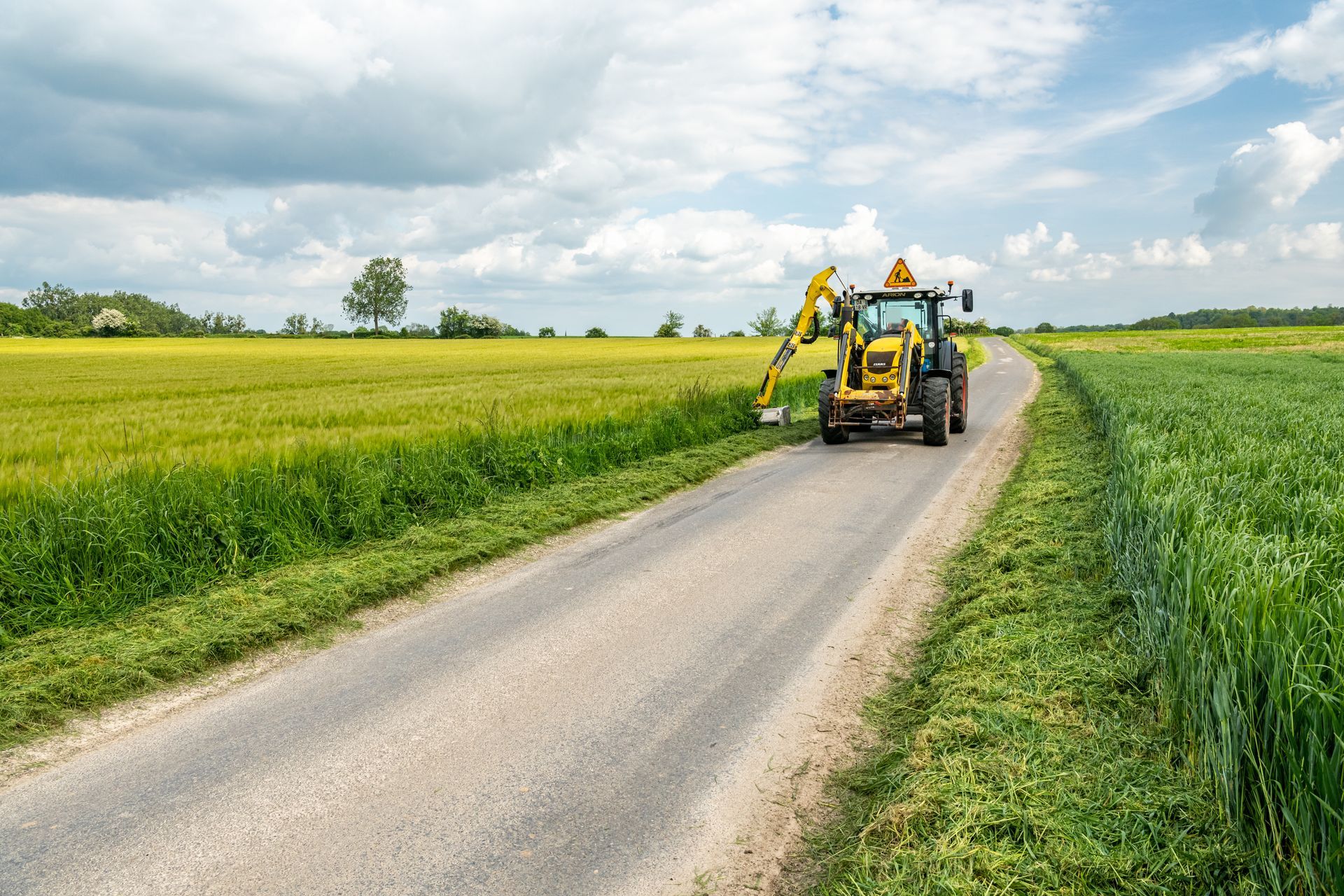 Tracteur jaune tondant l'herbe au bord d'une route de campagne.