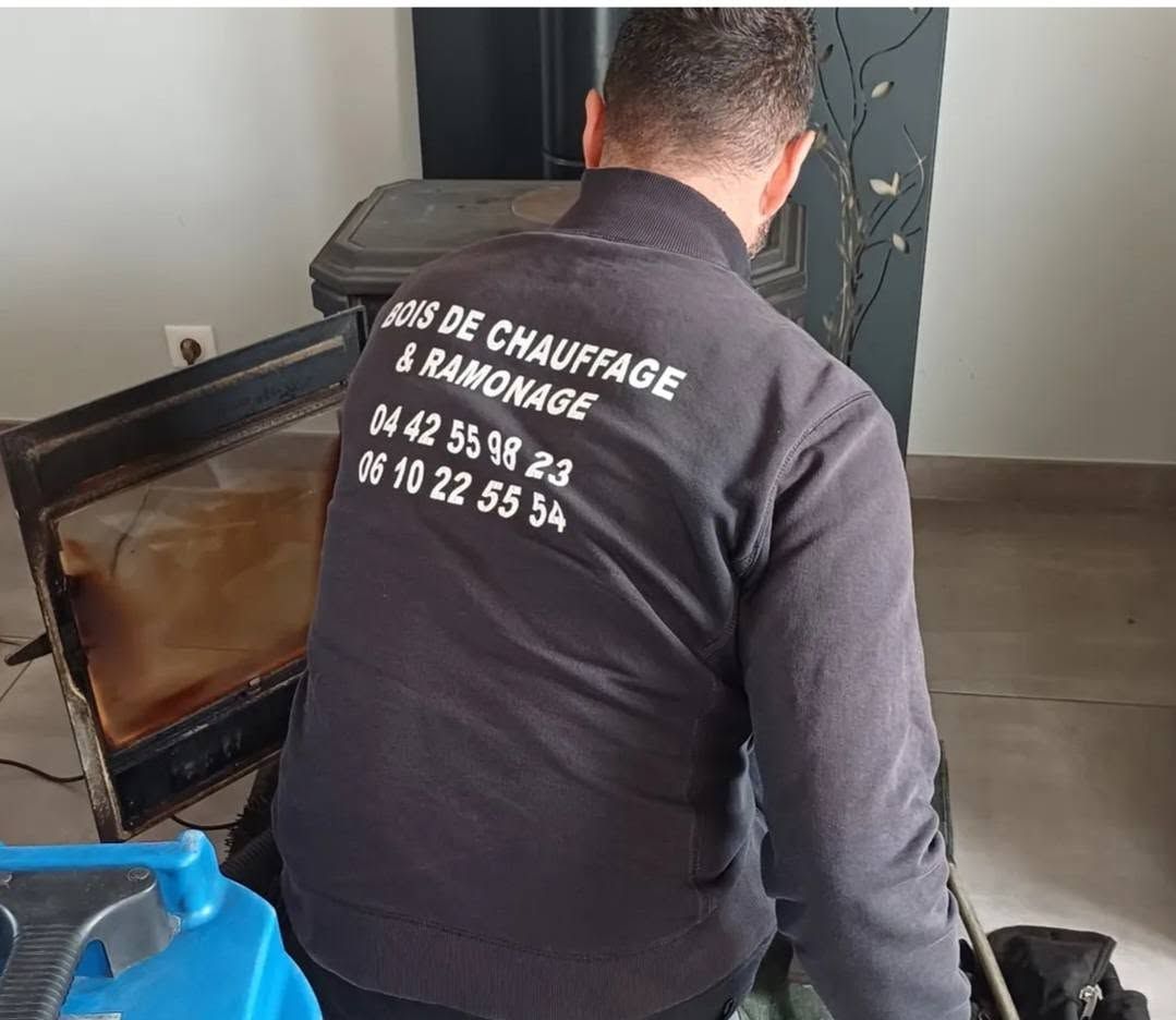 Un technicien en veste noire répare un poêle à bois à l'intérieur, près d'un conteneur bleu.