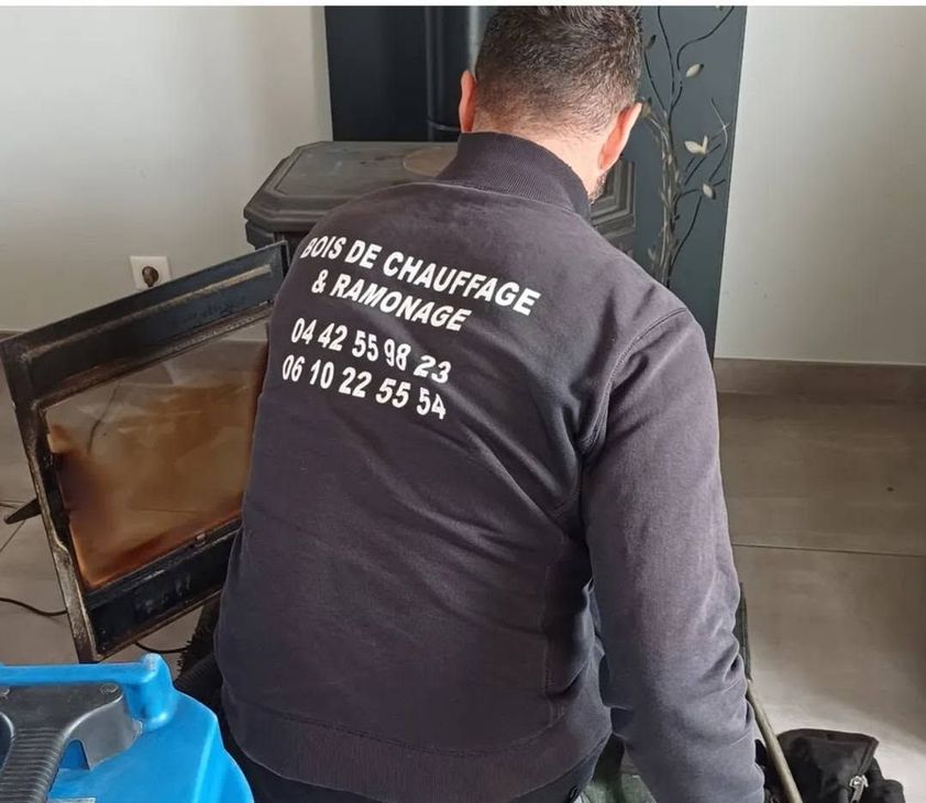Un technicien en veste noire répare un poêle à bois à l'intérieur, près d'un conteneur bleu.