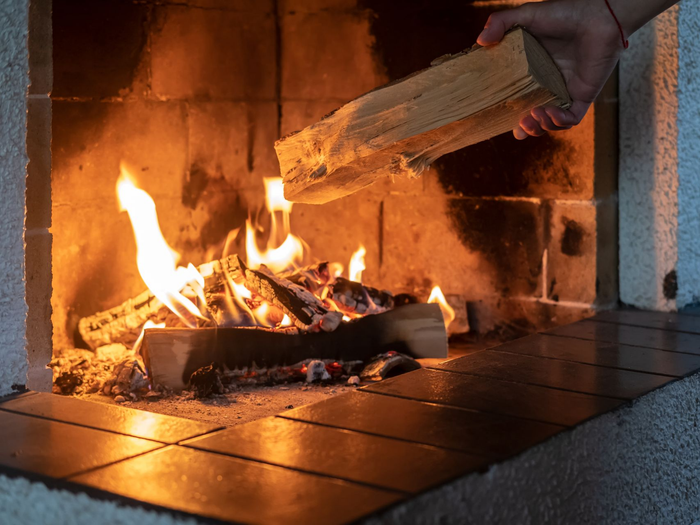 Une main dépose une bûche dans une cheminée allumée aux flammes orangées et entourée de briques.